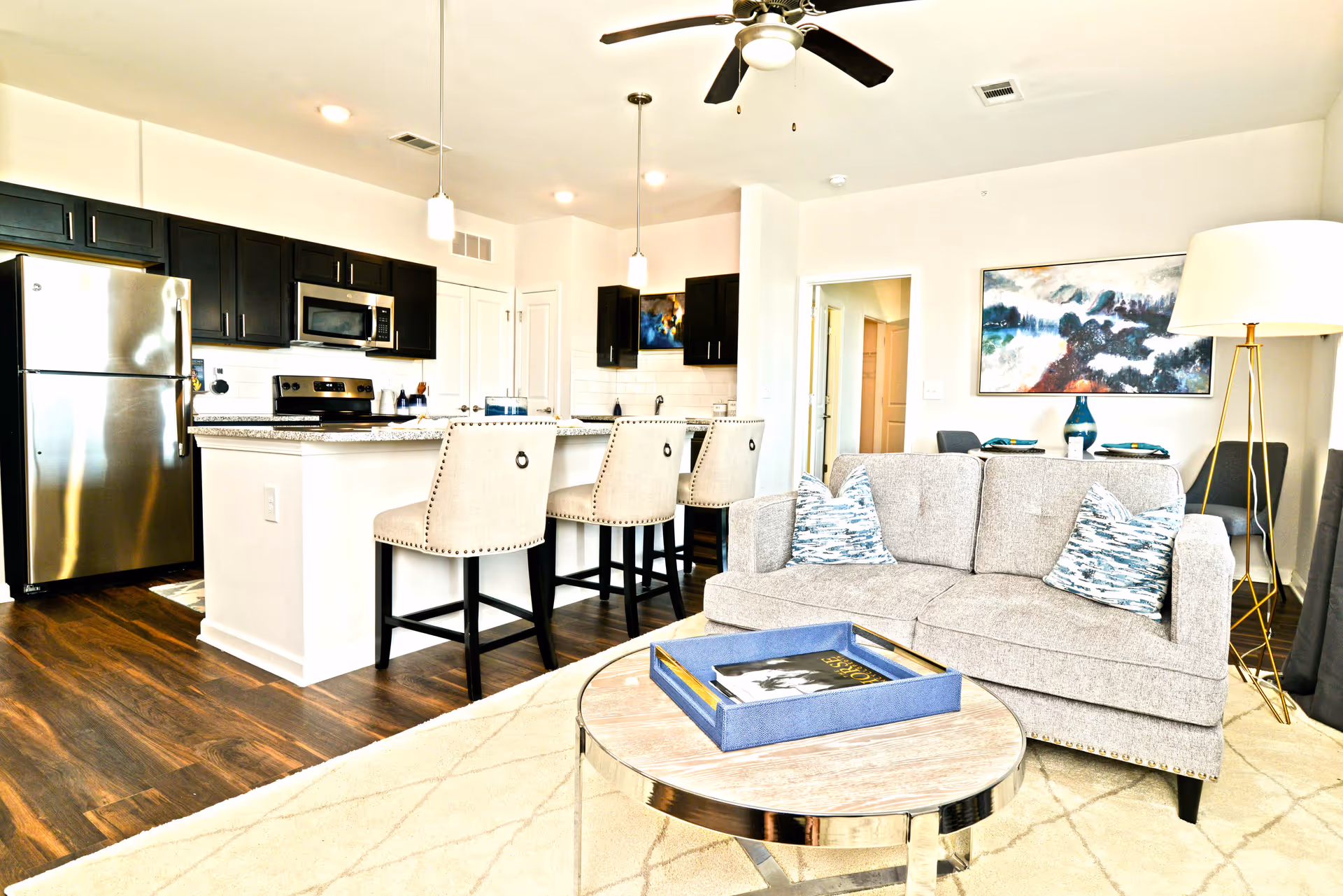 Bright open-plan living area showing a sofa and coffee table in front of a kitchen island with barstools and stainless steel appliances.