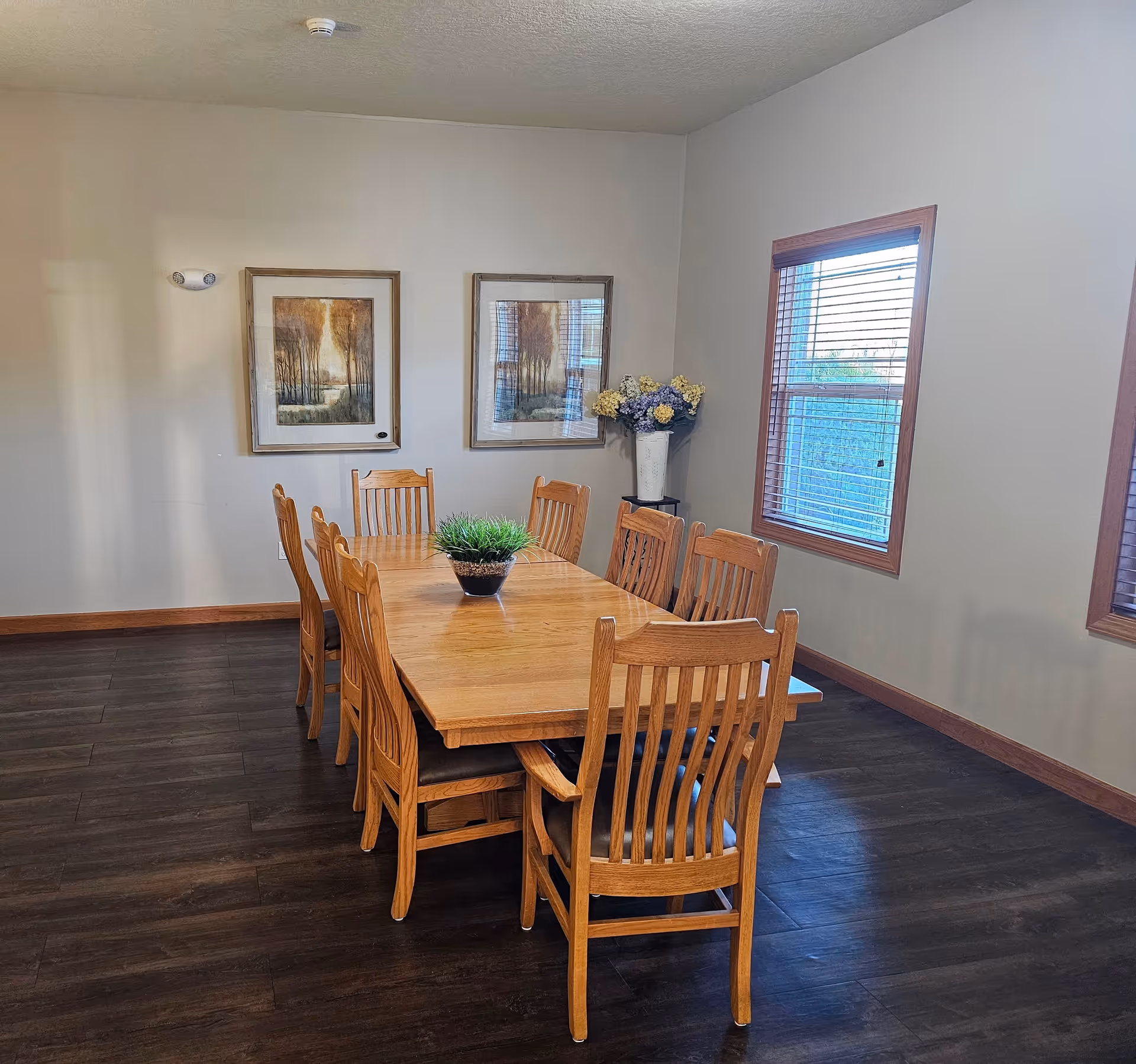 A dining room with a wooden table surrounded by eight matching wooden chairs. The table has a small green plant centerpiece. The room has two framed landscape paintings on the wall, a window with wooden blinds, and a vase with flowers on a small stand in the corner. The floor is dark wood, and the walls are light-colored.