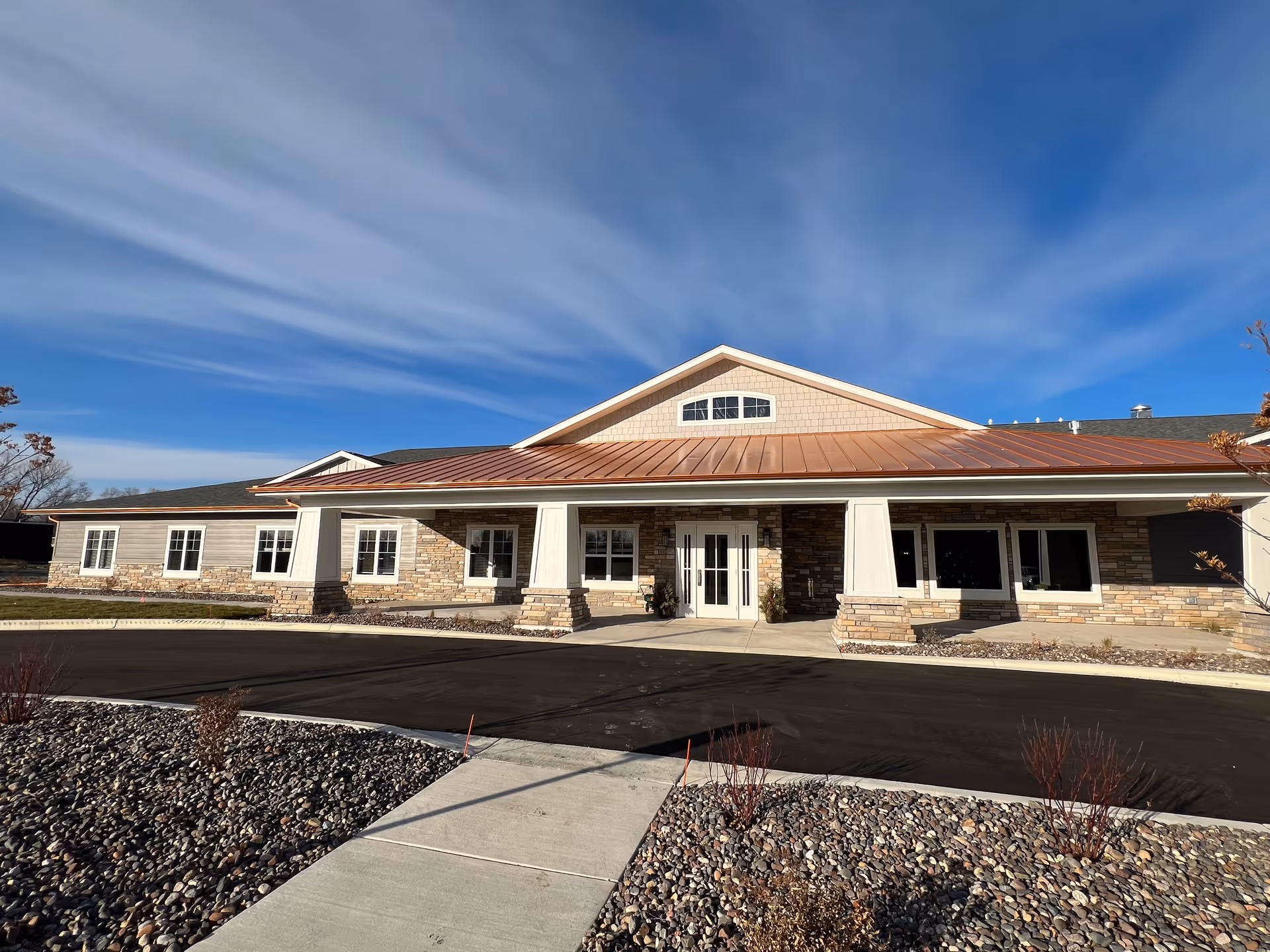 Front exterior view of a single-story building with a copper-colored metal roof, stone and siding facade, multiple windows, and a covered entrance supported by columns. The sky is clear with some wispy clouds, and there is a paved driveway and landscaped area with rocks and small shrubs in front.