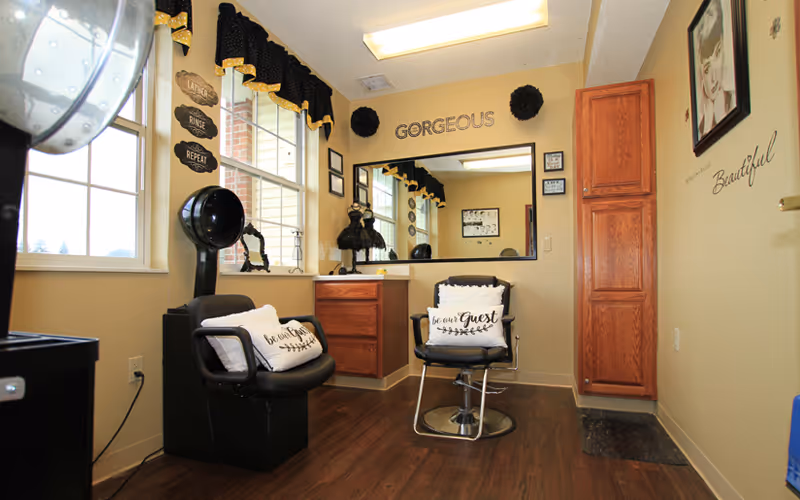 A cozy salon room with two black salon chairs, each with decorative pillows that say 'be our guest'. There is a large mirror on the wall with the word 'GORGEOUS' above it. The room has wooden flooring, a wooden cabinet, and windows with black valances. Various decorative wall signs and framed pictures adorn the walls.