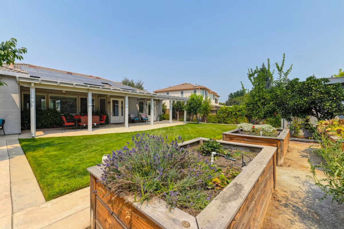 Backyard with a covered patio, lawn, seating area, and raised wooden garden beds under a clear sky.