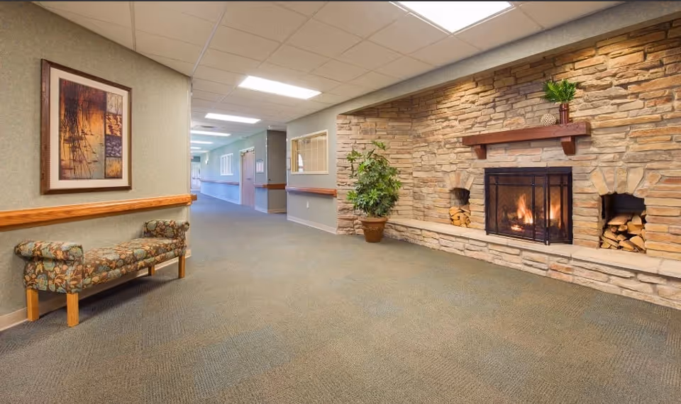 A hallway in a senior living facility featuring a stone fireplace with a fire burning, a potted plant, a floral upholstered bench, and a framed abstract painting on the wall. The hallway extends into the distance with doors and windows along the walls.