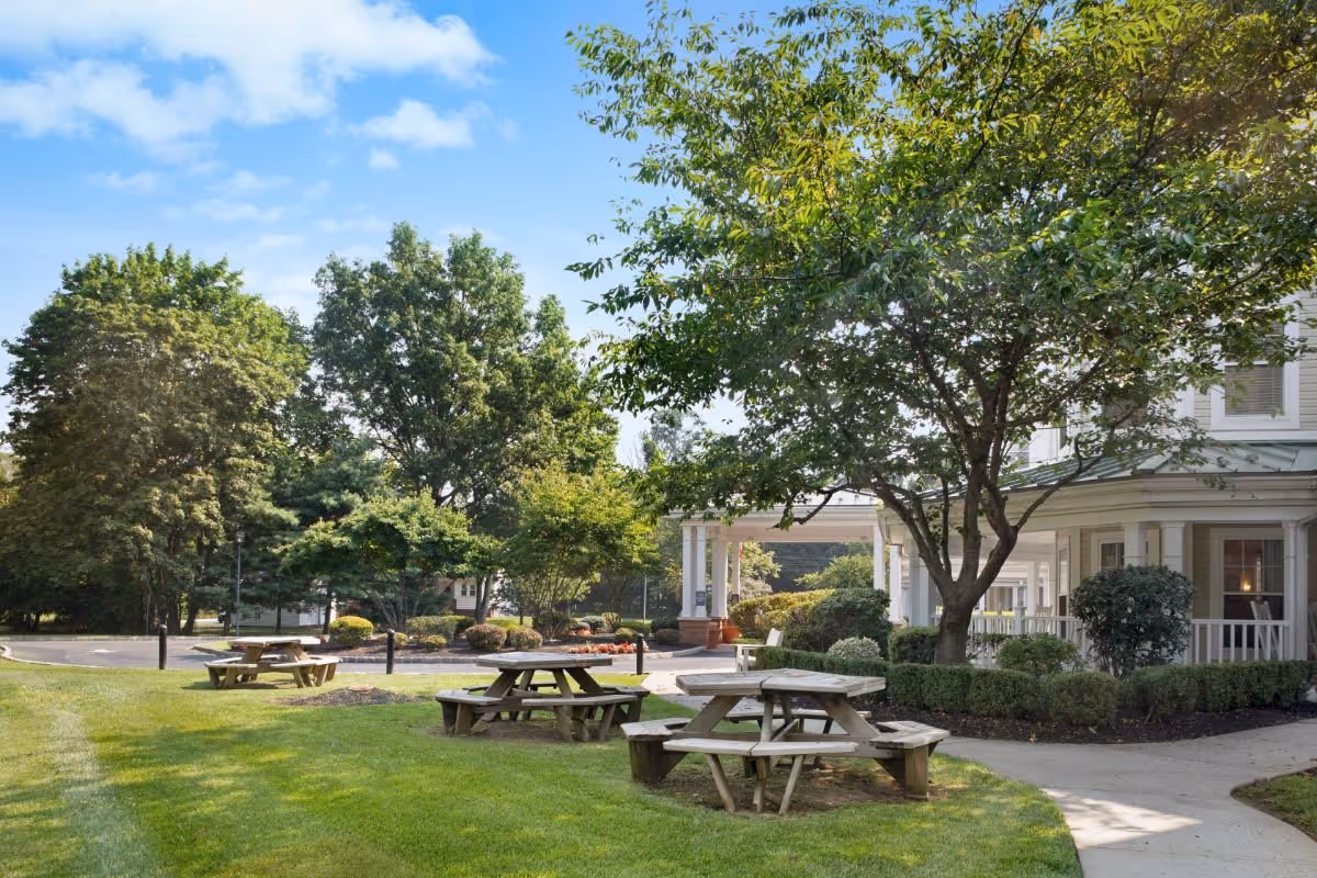 Outdoor area of a senior living facility with green grass, several wooden picnic tables, trees, and a building with a covered porch in the background under a blue sky with some clouds.