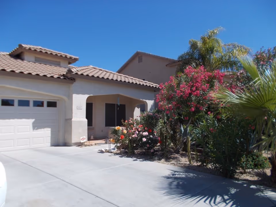 Exterior view of a single-story building with a tiled roof, a garage door, and a small covered porch. The front yard features a variety of flowering plants and palm trees under a clear blue sky.