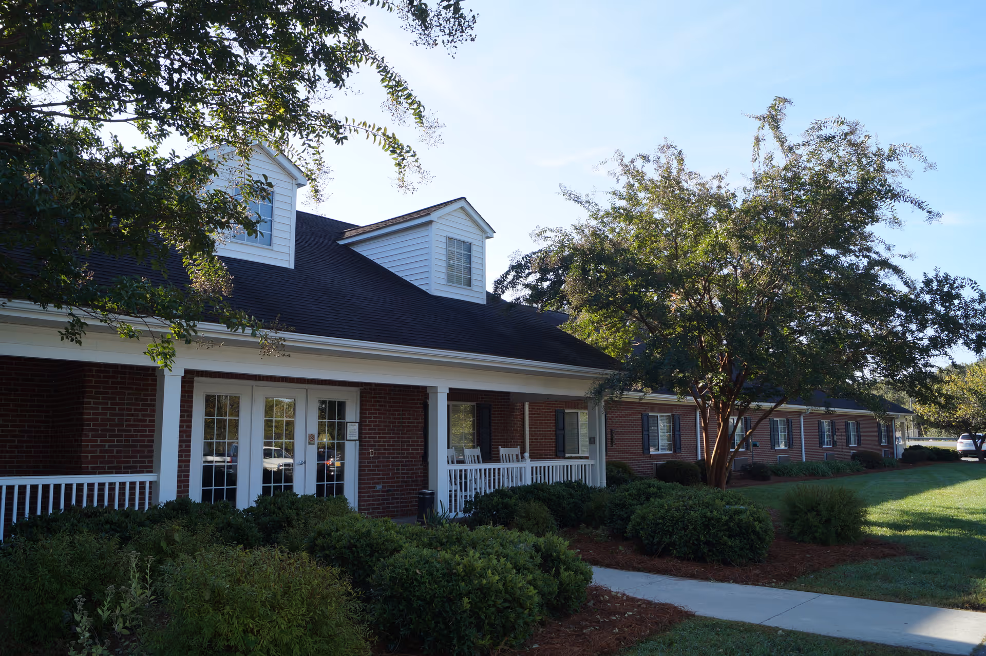 Brick single-story building front with a white-trimmed covered porch, rocking chairs, trees and landscaped shrubs on a sunny day.