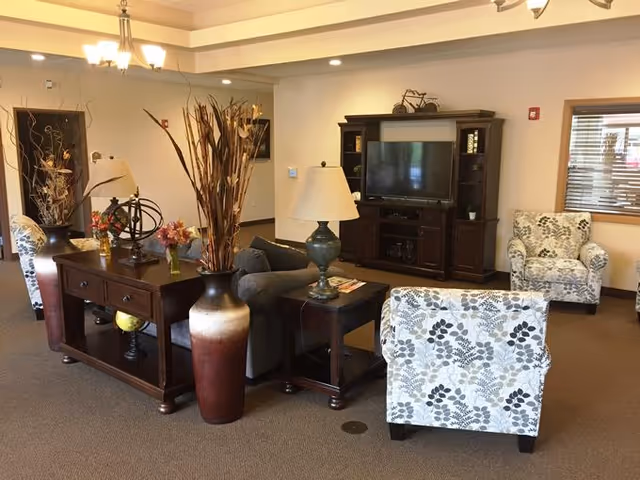 A cozy living room area in a senior living facility featuring a dark wood entertainment center with a TV, two patterned armchairs, a gray sofa, a wooden console table with decorative items including vases with dried plants and flowers, and a table lamp. The room has beige walls, carpeted floor, and a window with blinds.