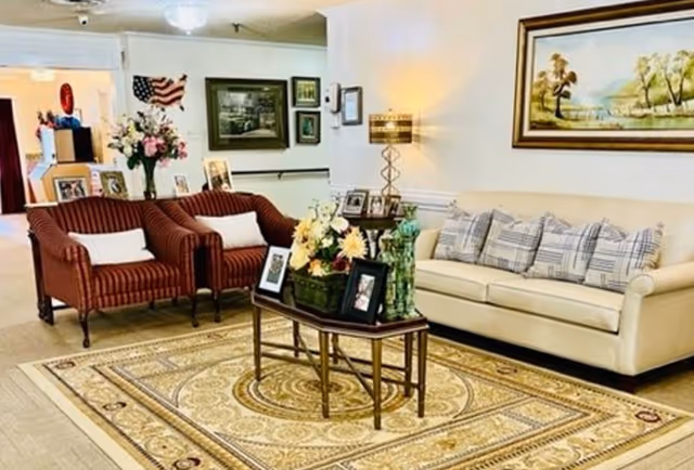 A cozy living room with a cream-colored sofa adorned with plaid pillows, two red striped armchairs with white cushions, a wooden coffee table with floral arrangements and framed photos, a patterned area rug, a standing lamp, and framed paintings on the walls.