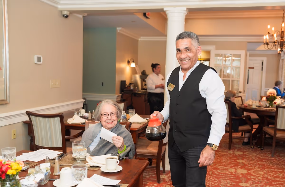 A smiling waiter holding a coffee pot stands next to an elderly woman seated at a dining table in a warmly lit dining room. The woman is holding a piece of paper and has a cup of coffee in front of her. Other elderly people are seated at tables in the background, and a staff member is visible near the back of the room.