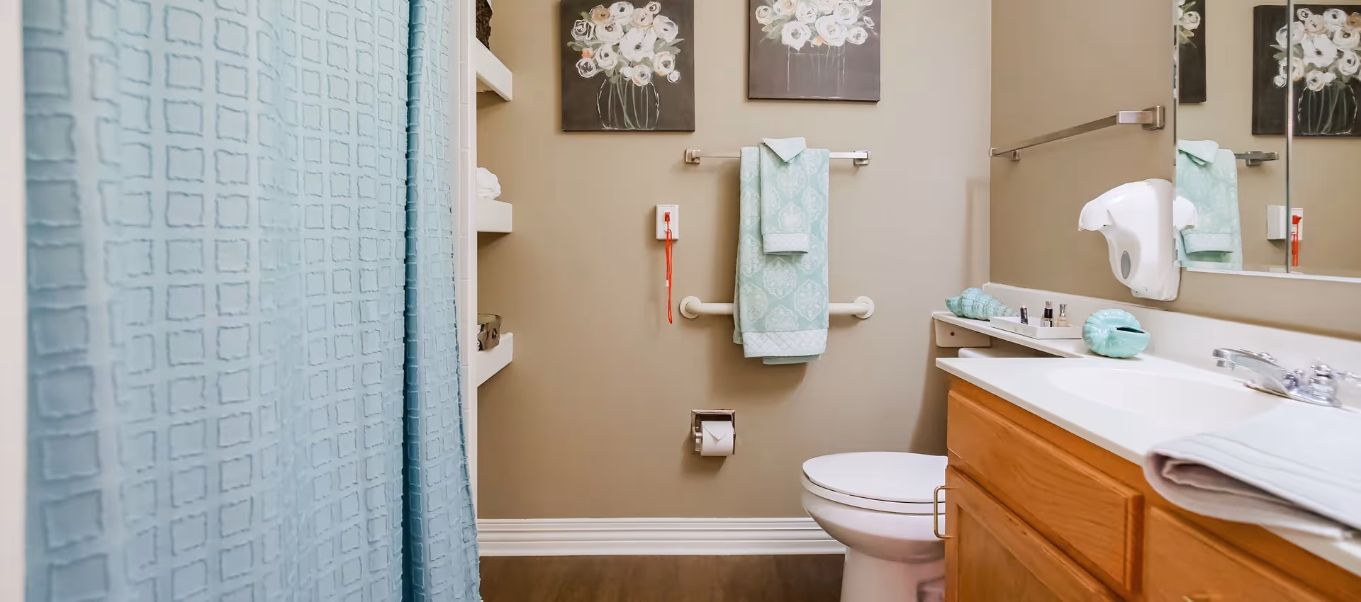 A clean and well-organized bathroom with a light blue shower curtain, a white toilet, a wooden vanity with a white countertop and sink, a wall-mounted soap dispenser, and two floral paintings above a towel rack holding light blue patterned towels.