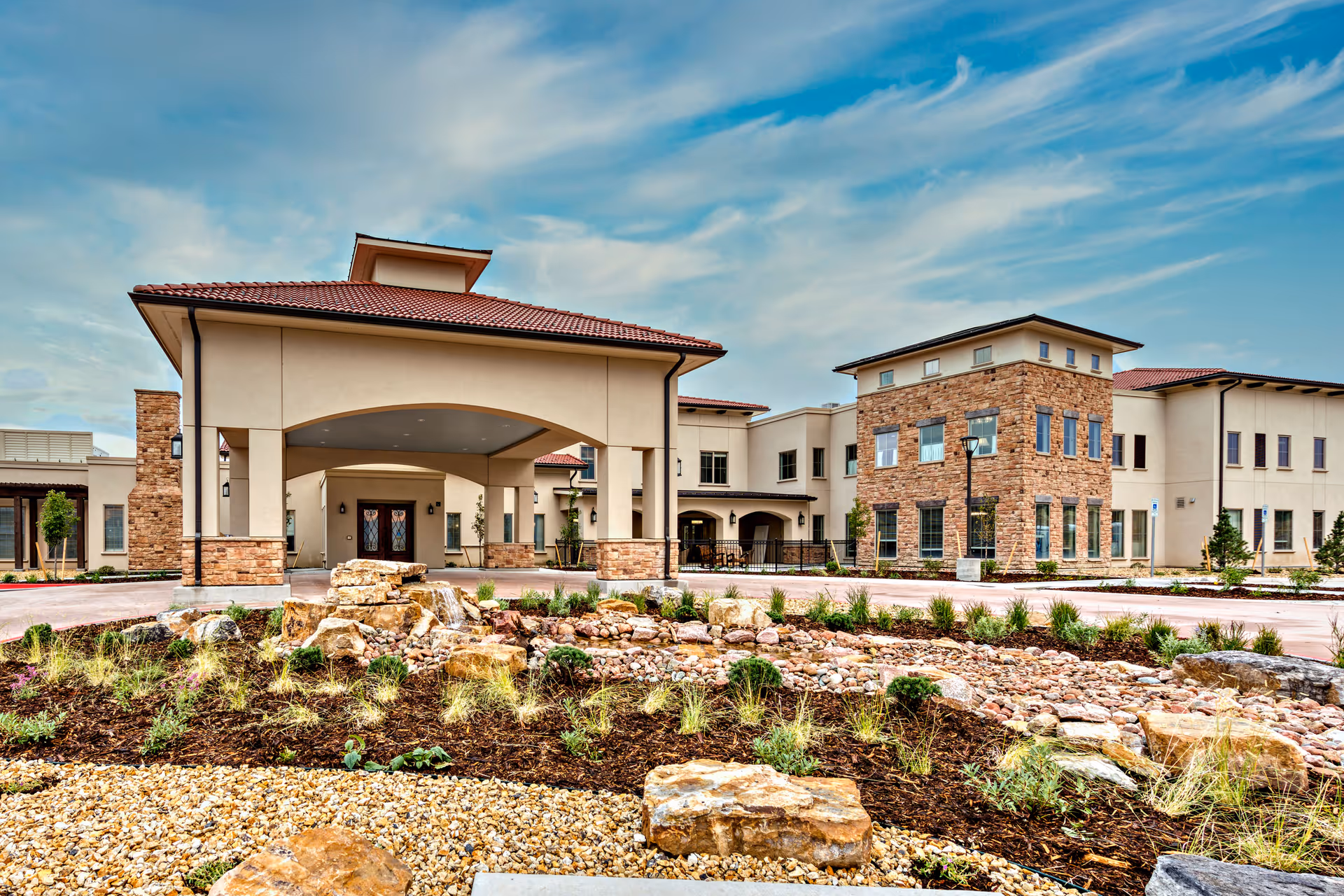 Exterior view of StoneCreek of Flying Horse Senior Living facility featuring a large entrance with a covered driveway, stone and stucco facade, landscaped garden with rocks and plants in the foreground, and a partly cloudy sky above.