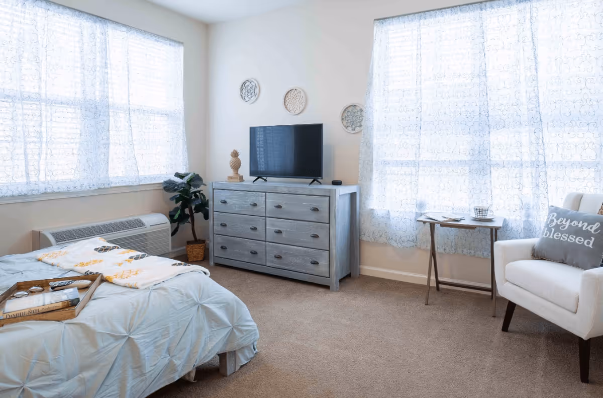 A bright bedroom with a bed covered in a light blue comforter and a tray holding books and glasses. There is a gray dresser with a flat-screen TV on top, a potted plant beside it, and three decorative wall hangings above. Two large windows with sheer patterned curtains let in natural light. A small side table with a cup and a book is next to a white armchair with a pillow that reads 'Beyond blessed.'