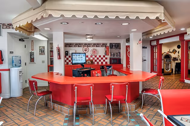 Interior view of a retro-style common area with a red and white color scheme, featuring a large red counter with several red chairs around it. The floor is tiled with a brown and orange pattern. In the background, there is a computer monitor on the counter, decorative plates on the wall, and a jukebox visible through a doorway with red-framed glass doors.