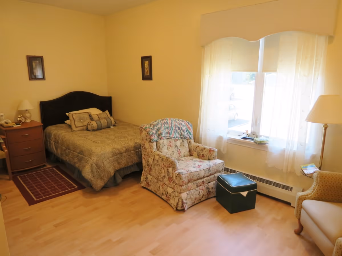 A cozy bedroom with a single bed covered in a patterned beige comforter and decorative pillows. Next to the bed is a wooden nightstand with a lamp, phone, and other small items. There is a floral upholstered armchair with a colorful knitted blanket draped over the back, a green ottoman in front of it, and another beige armchair partially visible on the right. A large window with sheer white curtains lets in natural light, and the floor is light wood.