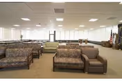 A spacious room with multiple rows of patterned sofas and armchairs arranged facing forward. The room has large windows covered with white blinds, a beige carpeted floor, and a white ceiling with recessed lighting. There are a few pieces of furniture and decorations along the walls.