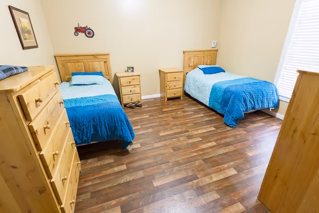 A bright bedroom with two single beds, each with blue bedding and wooden headboards. Between the beds are two wooden nightstands with small decorative items. There is a wooden dresser on the left side and another wooden piece of furniture on the right. The room has wood flooring and a window with blinds on the right wall. A framed picture and a small red tractor decoration hang on the beige walls.