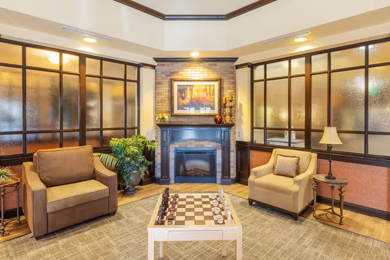 A cozy sitting area with two armchairs facing a chessboard on a small table. Behind the chessboard is a fireplace with a framed painting above it and decorative candles on the mantel. The room has frosted glass panels on the walls and a carpeted floor.