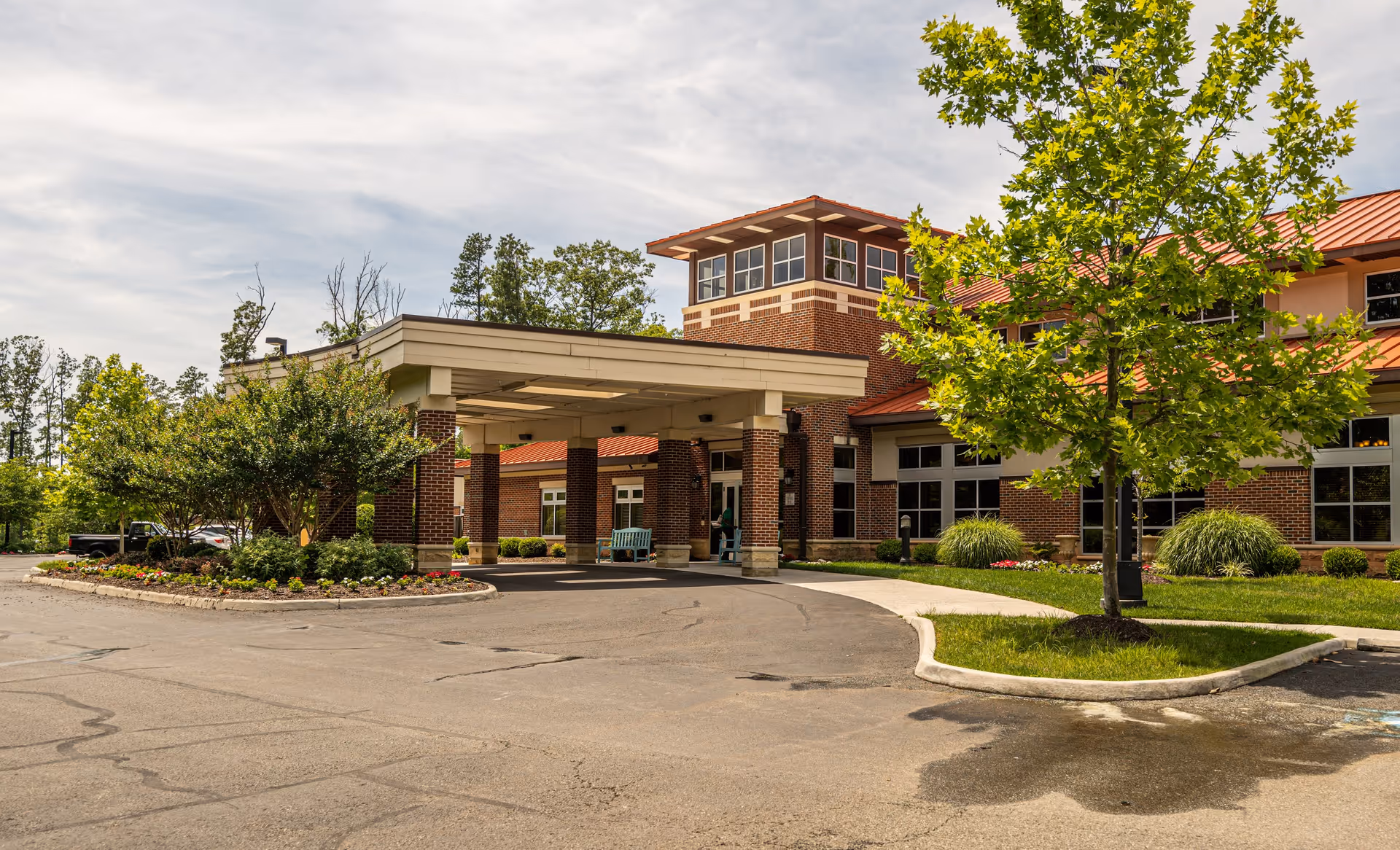Exterior view of Arden Courts Richmond facility showing a brick building with a covered entrance, surrounded by landscaped greenery and trees under a partly cloudy sky.