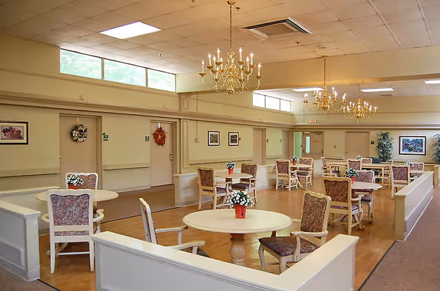 A spacious dining room with multiple round tables and chairs arranged neatly. Each table has a small flower pot with white flowers. The room features beige walls, several framed pictures, decorative wreaths on doors, large windows near the ceiling, and elegant chandeliers hanging from the ceiling.