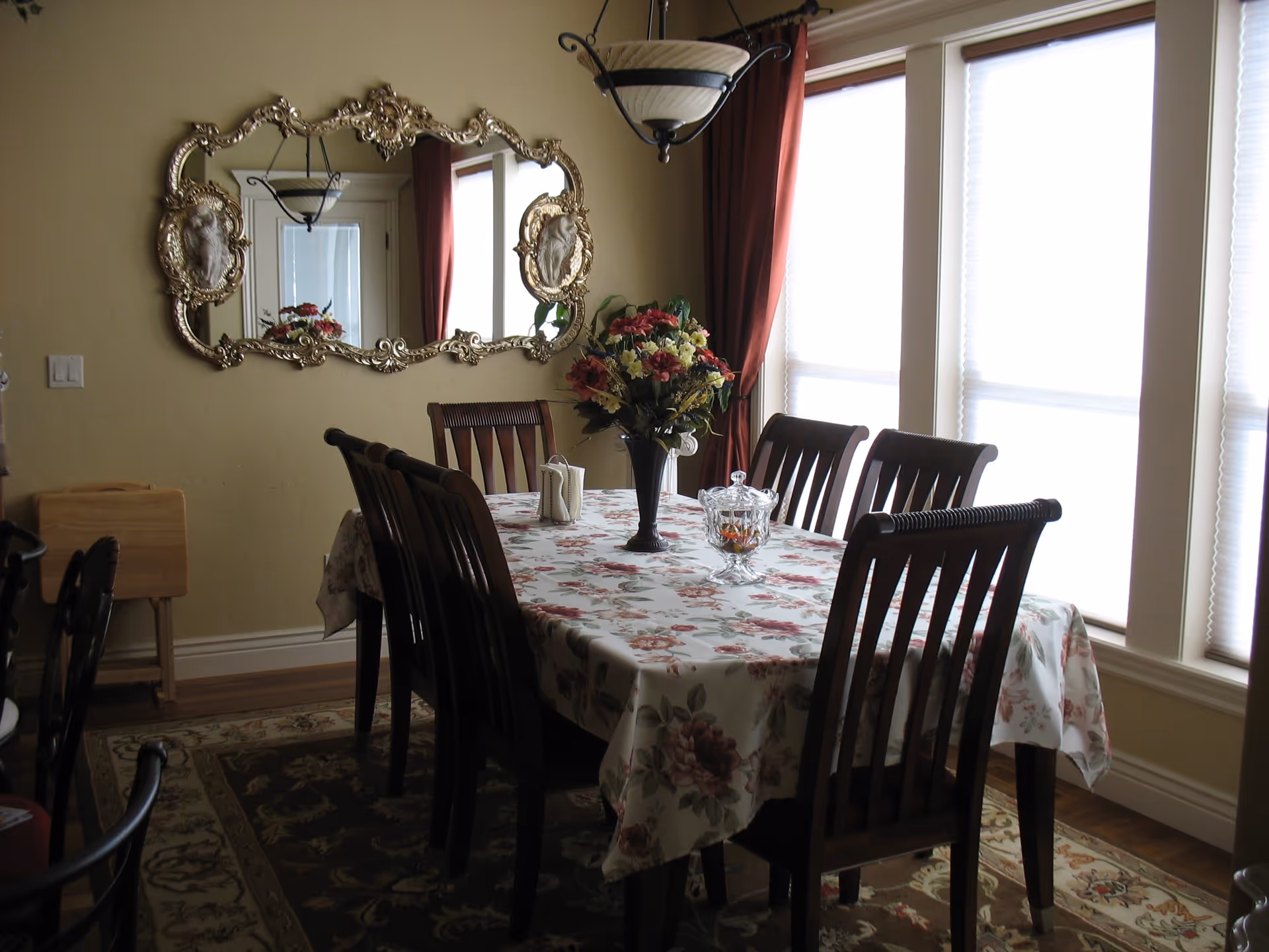 A dining room with a rectangular wooden table covered with a floral tablecloth. Six wooden chairs surround the table. A vase with a colorful flower arrangement and a glass candy dish are placed on the table. The room has large windows with white blinds and red curtains. An ornate gold-framed mirror hangs on the beige wall, reflecting part of the room and a ceiling light fixture.