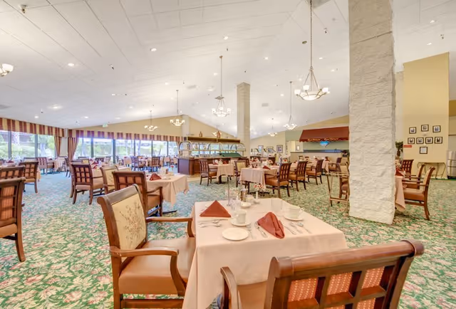Spacious dining room with multiple tables covered in light pink tablecloths, set with cups, plates, and folded napkins. The room features large windows with striped curtains allowing natural light, green patterned carpet, and several chandeliers hanging from a high ceiling. Wooden chairs with cushioned seats surround the tables, and a buffet area is visible in the background.