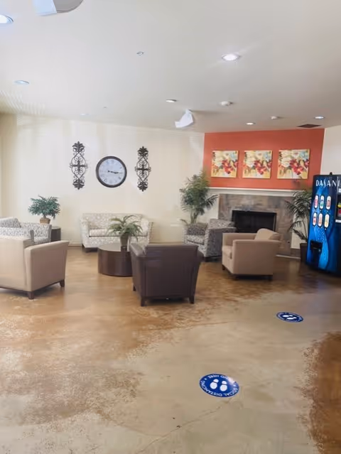 A cozy living room area with several armchairs and a small sofa arranged around a central coffee table. The room features a fireplace with three floral paintings above it, a large clock on the wall, decorative wall hangings, and potted plants. There is a blue Dasani vending machine on the right side and social distancing floor markers on the polished concrete floor.