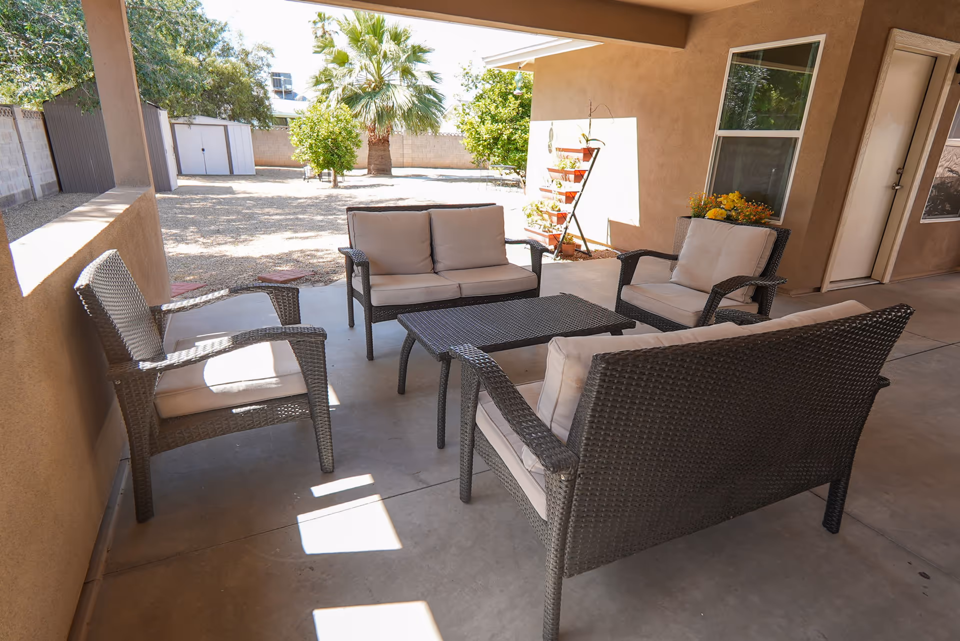 Outdoor covered patio area with wicker furniture including two armchairs, two loveseats, and a rectangular coffee table. The patio overlooks a yard with gravel ground, a palm tree, and other greenery. There is a vertical plant stand with potted plants against the wall near a window and door.