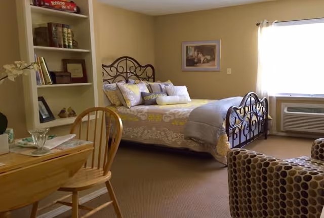 A cozy bedroom in Grayson Court featuring a bed with an ornate metal headboard and footboard, decorated with multiple pillows and a patterned bedspread. To the left, there is a wooden table with a chair and a built-in bookshelf filled with books and decorative items. A patterned armchair is partially visible in the foreground, and a window with white curtains allows natural light to brighten the room. An air conditioning unit is installed below the window.