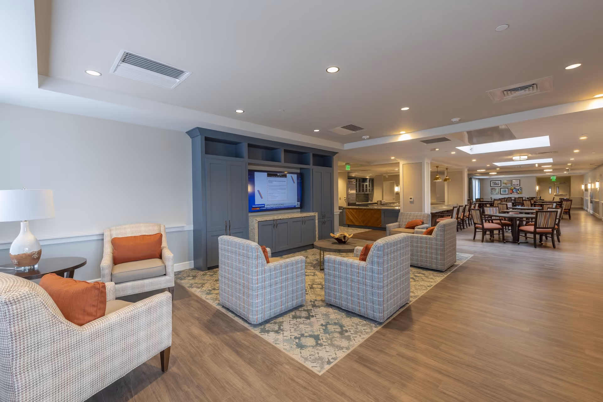 A spacious senior living common area featuring a seating arrangement with four plaid armchairs and two beige armchairs with orange cushions around a round coffee table on a patterned rug. A large built-in entertainment center with a TV is against the wall. In the background, there is a dining area with multiple tables and chairs, and a kitchen area with pendant lighting. The room has wood flooring, recessed lighting, and a neutral color palette.