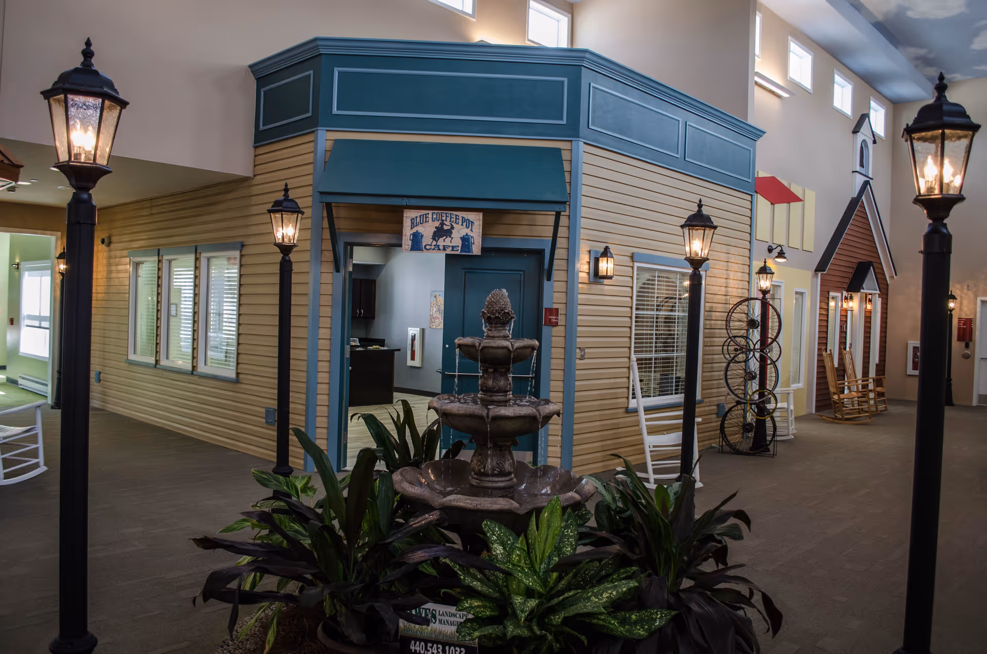 Indoor area designed to look like a small town street with building facades, including a cafe named Blue Coffee Pot Cafe. There are street lamps, a three-tiered water fountain surrounded by plants, rocking chairs, and decorative elements like a bicycle wheel sculpture. The ceiling is painted with clouds.