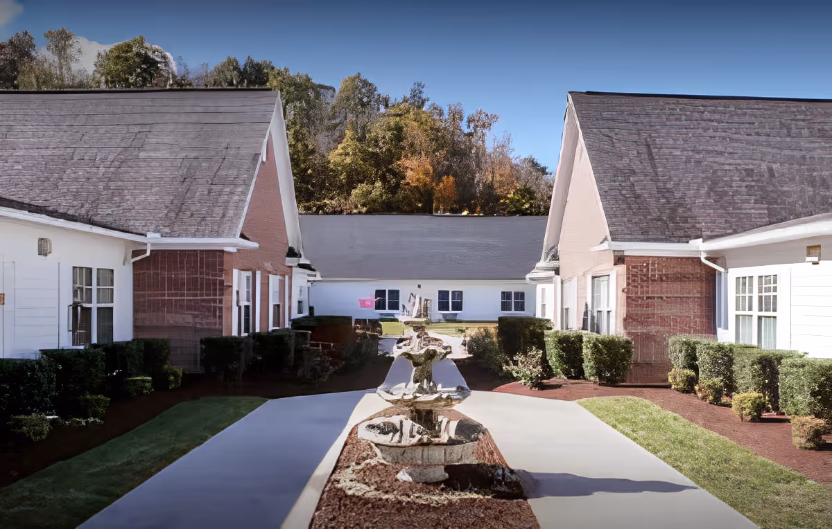 Outdoor courtyard area of a senior living facility with a central pathway flanked by trimmed bushes and a decorative fountain in the middle. The buildings on either side have white and brick exteriors with multiple windows, and trees are visible in the background under a clear blue sky.