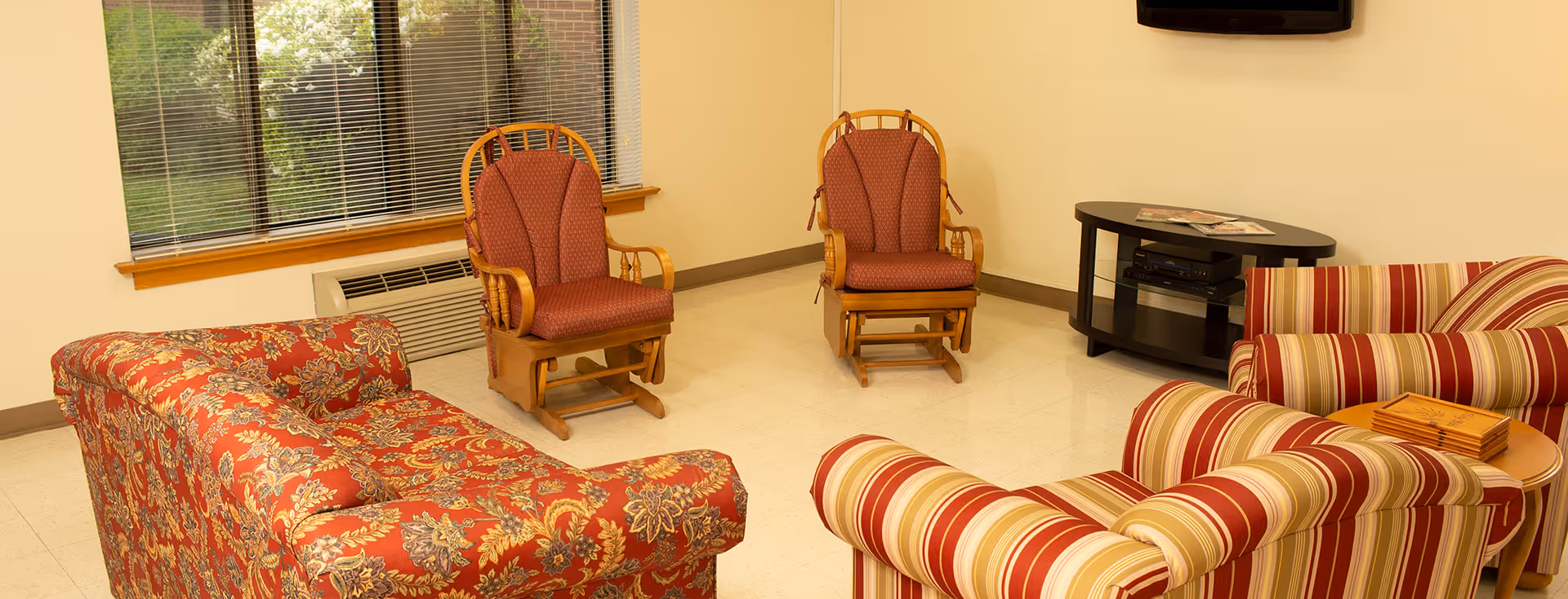 A cozy living room area with patterned upholstered furniture including a floral sofa, two striped armchairs, and two wooden rocking chairs with red cushions. There is a window with blinds, a small black TV stand with a flat-screen TV mounted on the wall above it, and a small round wooden table with a wooden box on it.