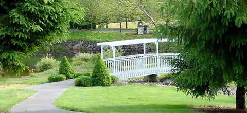 A white pedestrian bridge crosses a small creek in a manicured outdoor park setting.