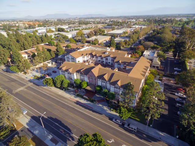 Aerial view of a multi-story, Tudor-style assisted living building along a tree-lined street with surrounding parking and neighborhood.