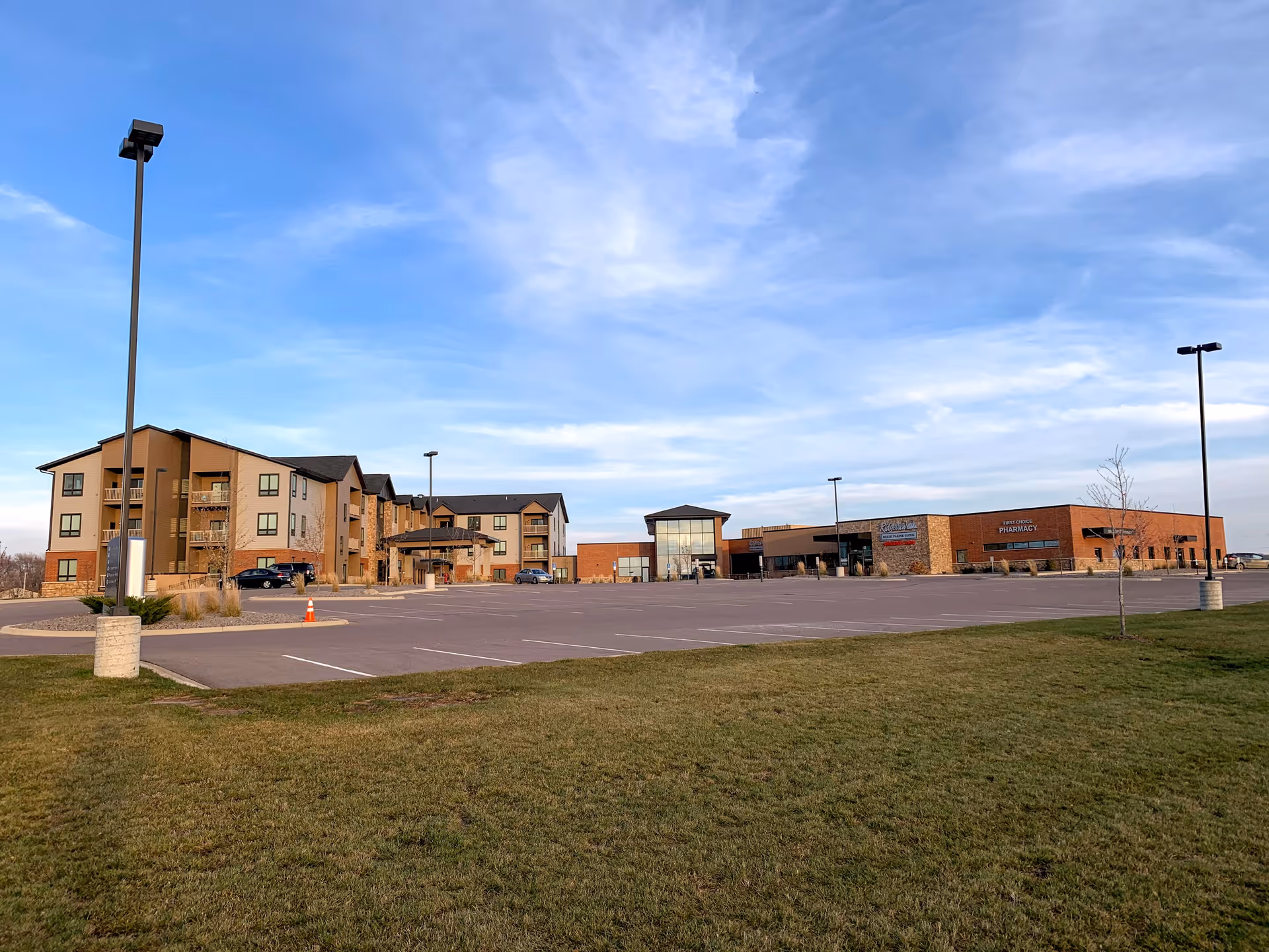 Wide exterior view of a senior living facility with a large parking lot in front, a multi-story residential building on the left, and a single-story commercial building on the right under a partly cloudy sky.