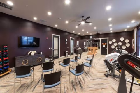 A spacious fitness and activity room with several chairs arranged in rows facing a wall-mounted TV. The room has dark walls, a light wood-patterned floor, ceiling fans, and recessed lighting. On the right side, there is exercise equipment including a treadmill and weights. Decorative plates are displayed on one wall, and there is a console table with flowers beneath the TV.