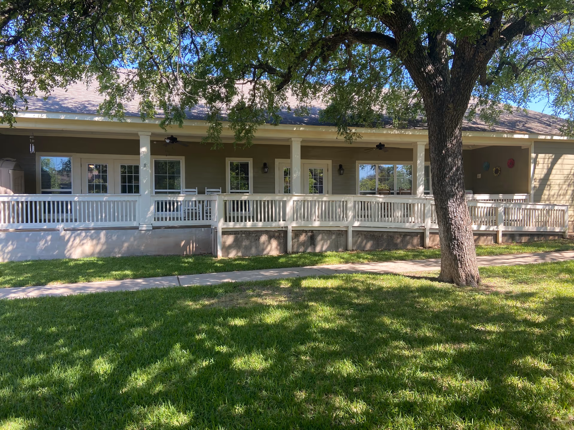 Exterior view of Mercy House Waco showing a single-story building with a covered porch, white railing, and several windows and doors. There is a large tree in the foreground casting shadows on the green grass and a sidewalk running parallel to the porch.