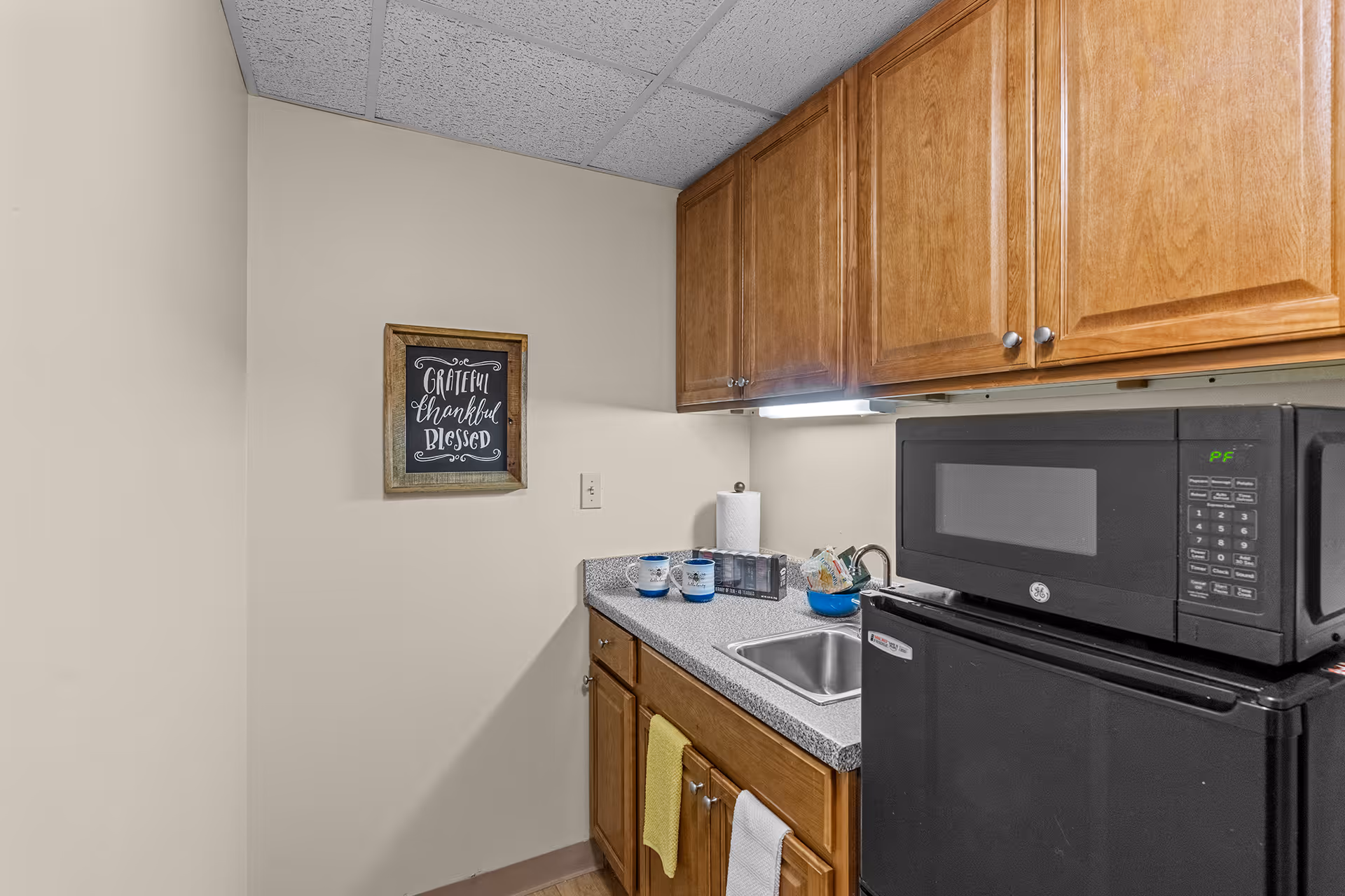 Small kitchenette with wooden cabinets, a countertop with a sink, a black microwave on top of a black mini refrigerator, and a wall decoration that reads 'Grateful Thankful Blessed'.