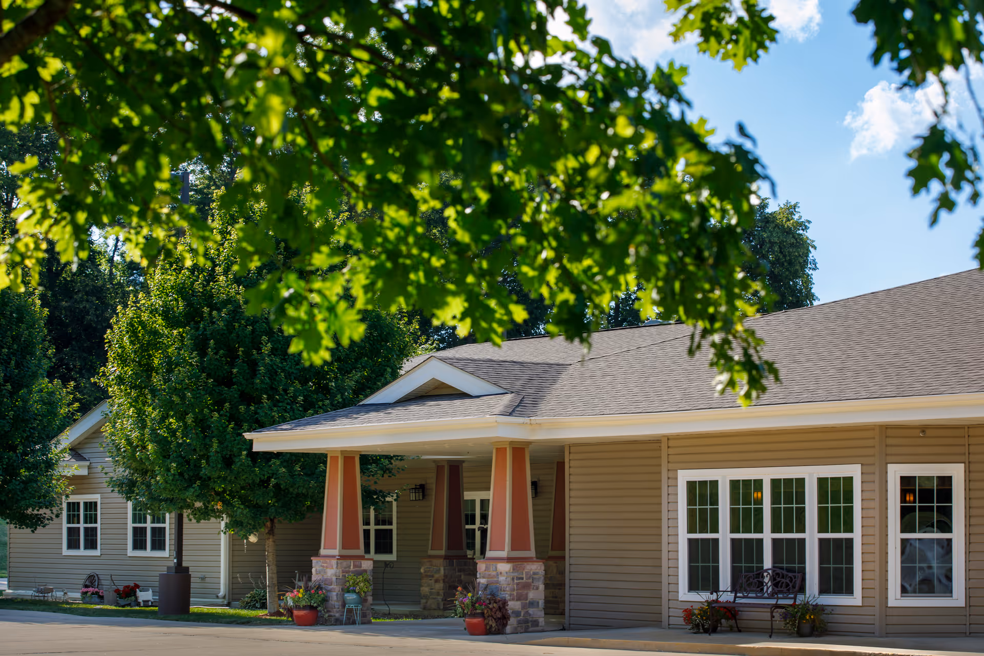 Front exterior of a single-story assisted living building with a covered entrance, columns, windows, and trees in the foreground.