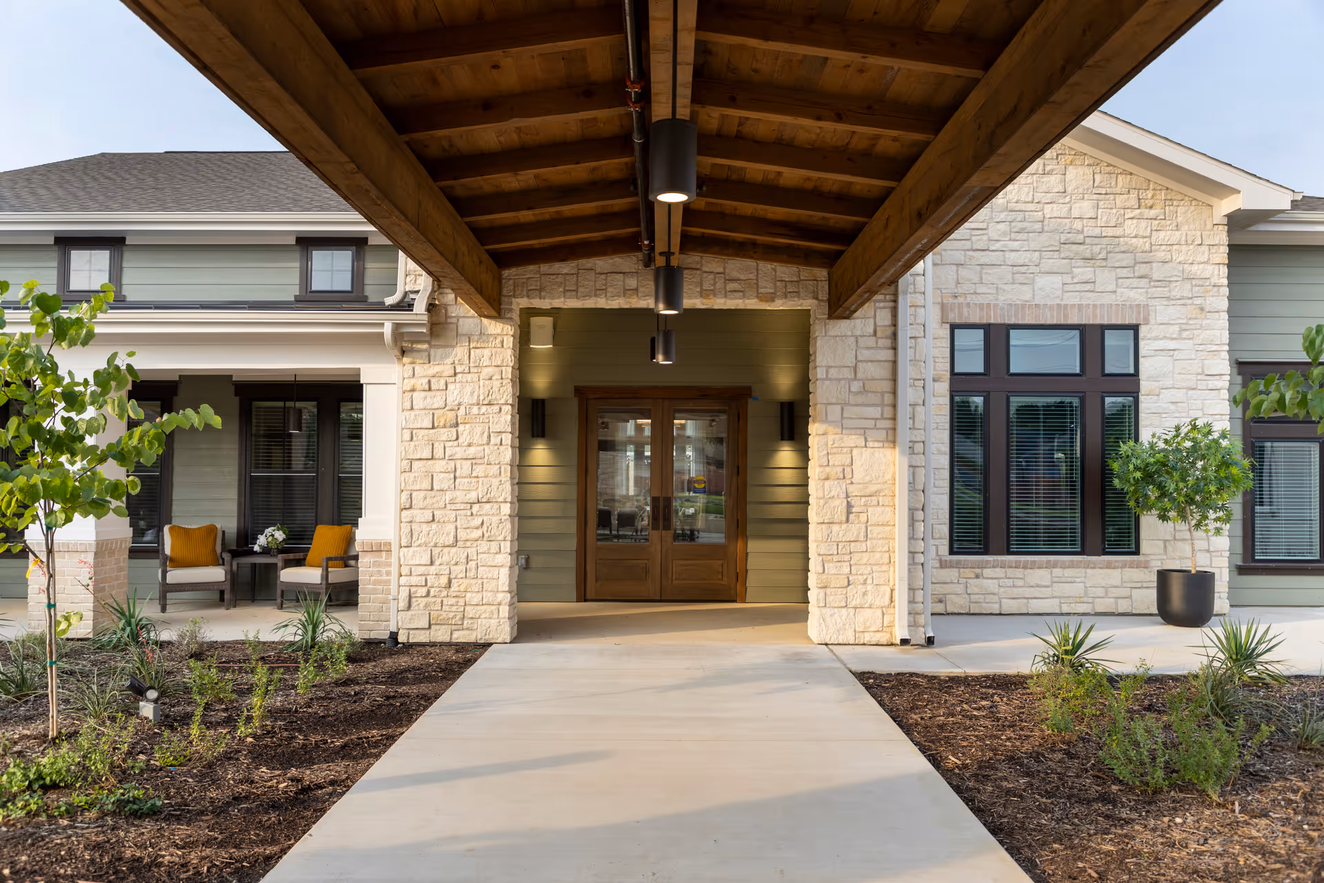 Entrance to a senior living facility with a covered walkway featuring a wooden ceiling, stone pillars, and double wooden doors. There are small landscaped garden beds with plants on either side of the concrete path leading to the entrance. To the left, there is a seating area with two chairs and a small table with a flower arrangement.