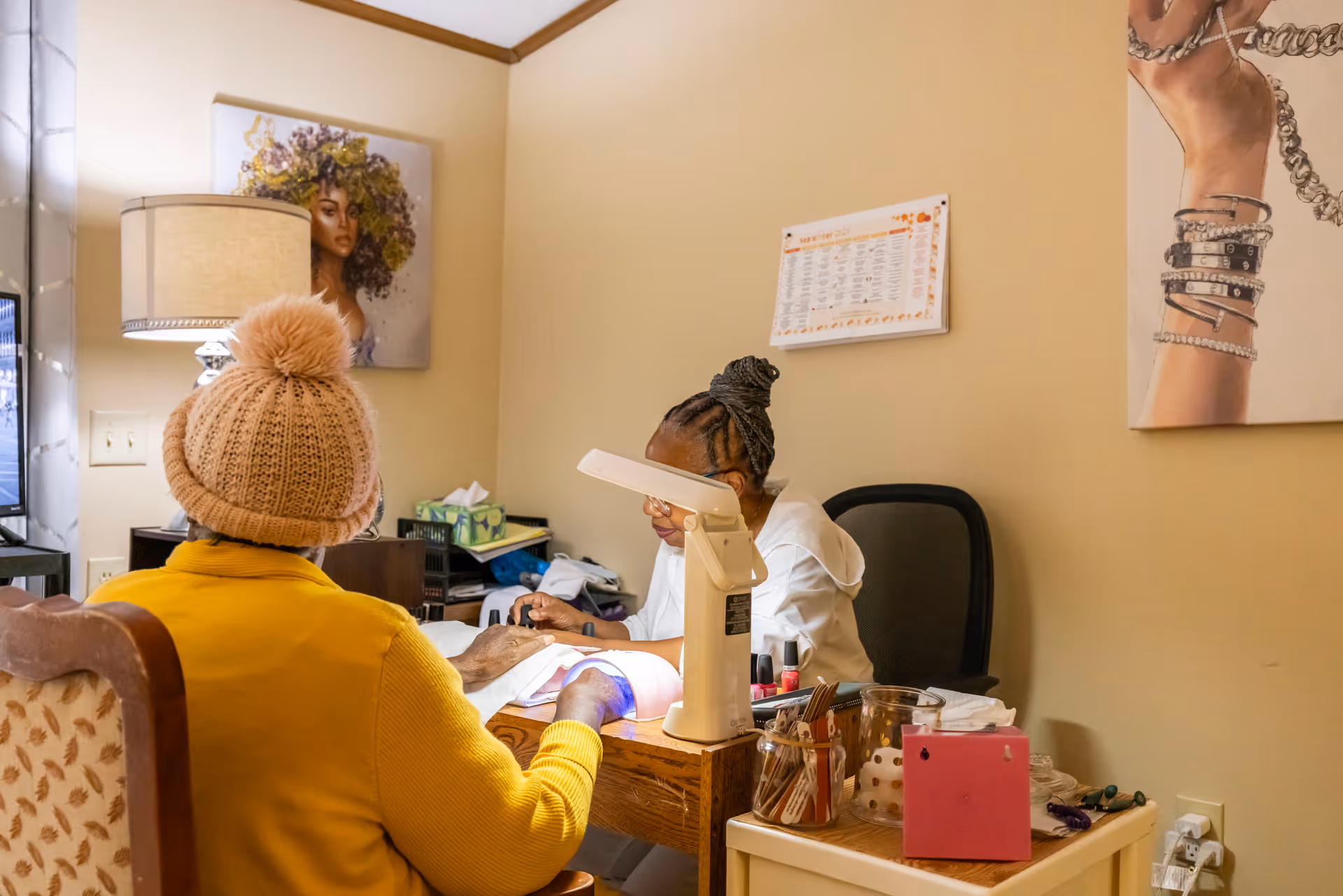 A person wearing a knitted hat and yellow sweater is sitting at a table receiving a manicure from another person in a cozy room with beige walls. The table has nail polish bottles and manicure tools, and the room is decorated with artwork and a lamp.