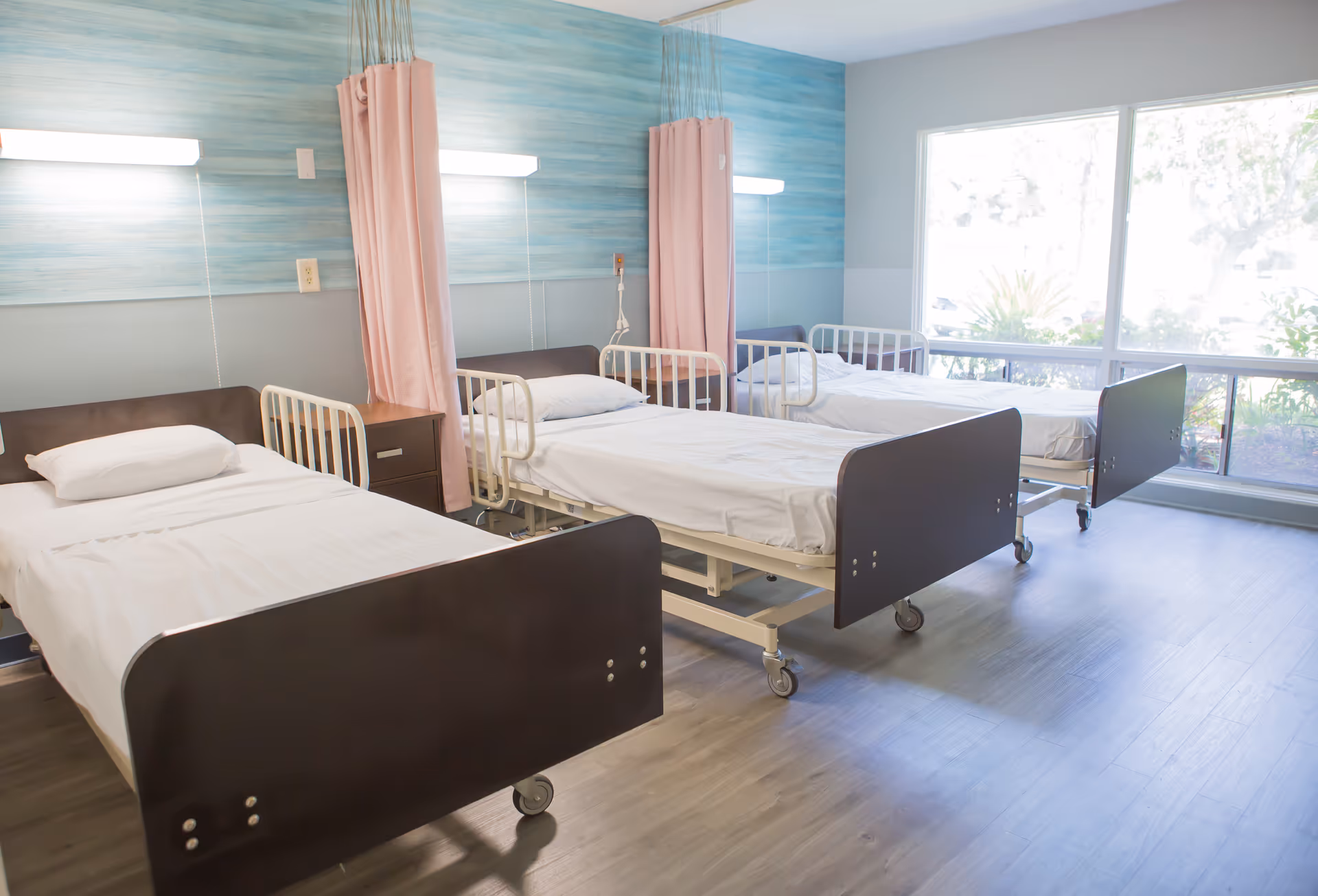 A bright hospital or healthcare center room with three single beds lined up side by side, each with white sheets and pillows. The beds have dark brown headboards and footboards, and are equipped with side rails. Pink privacy curtains hang between the beds. There is a large window on the right side letting in natural light, and the walls have a blue and gray color scheme.