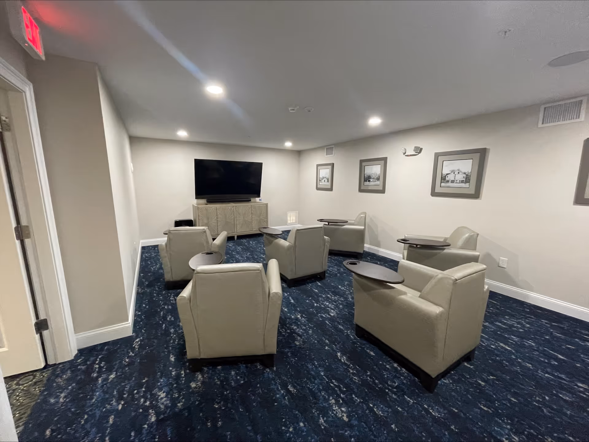A small lounge area with six beige armchairs arranged in a semi-circle facing a large flat-screen TV mounted on a cabinet. The room has blue patterned carpet, light-colored walls with framed pictures, and recessed ceiling lights.