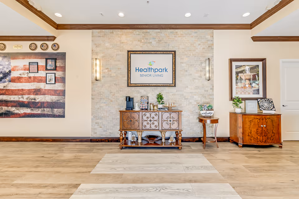Interior view of a senior living facility lobby area with a wooden sideboard against a light stone accent wall. Above the sideboard is a framed sign that reads 'Healthpark Senior Living'. The sideboard holds a coffee maker, cups, and decorative plants. To the left, there is a large American flag wall art with framed pictures and military emblems above it. To the right, there is a small round table with a welcome sign and a wooden cabinet with framed artwork and decorative items on top. The floor is light wood with a patterned rug area in front of the sideboard.