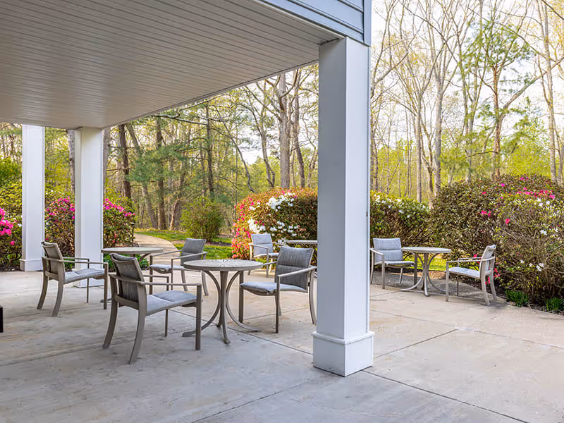 Outdoor covered patio area with several round tables and chairs arranged for seating, surrounded by bushes with pink and white flowers and trees in the background.
