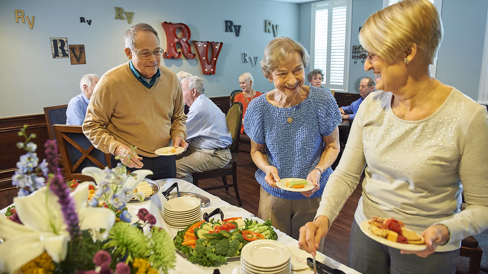 Three elderly people smiling and serving themselves food from a buffet table in a dining room decorated with various 'Rv' letters on the wall. Other elderly people are seated and conversing in the background.