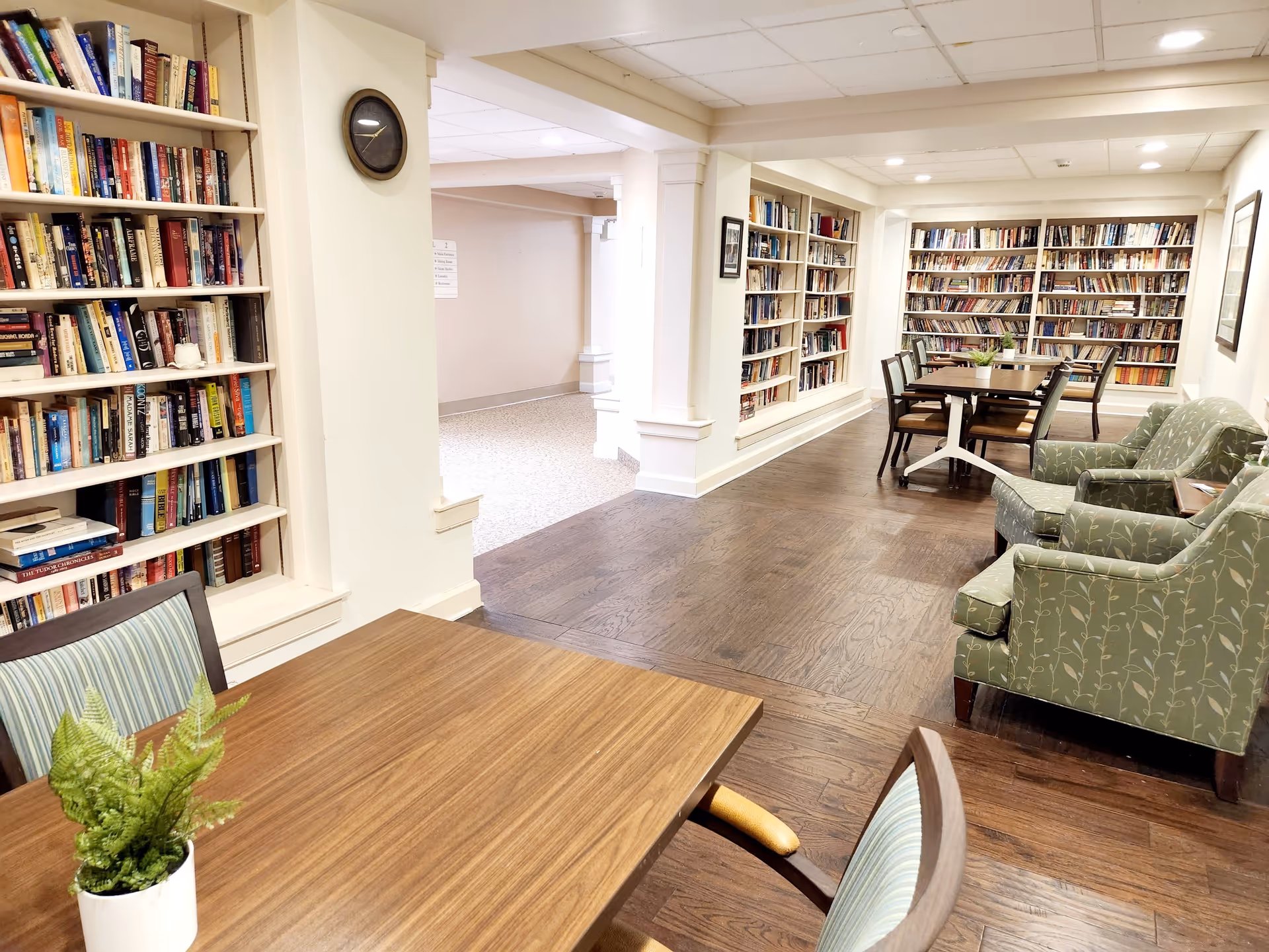 A cozy library room with wooden floors, bookshelves filled with books lining the walls, several tables with chairs, and two green upholstered armchairs. A small potted plant is on the table in the foreground, and a clock is mounted on a white wall near the entrance to another room.