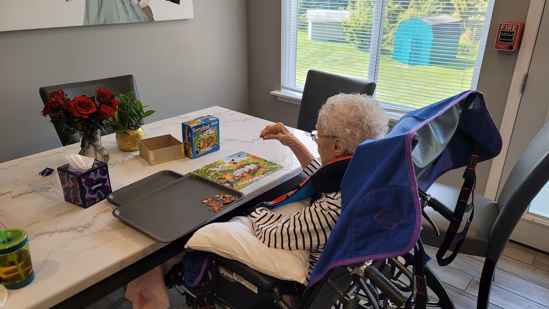 An elderly woman in a wheelchair is sitting at a marble table working on a jigsaw puzzle. The table has puzzle pieces, a puzzle box, a tissue box, a vase with red roses, and a small green plant. The room has a window with blinds and a door with a fire alarm on the wall nearby.