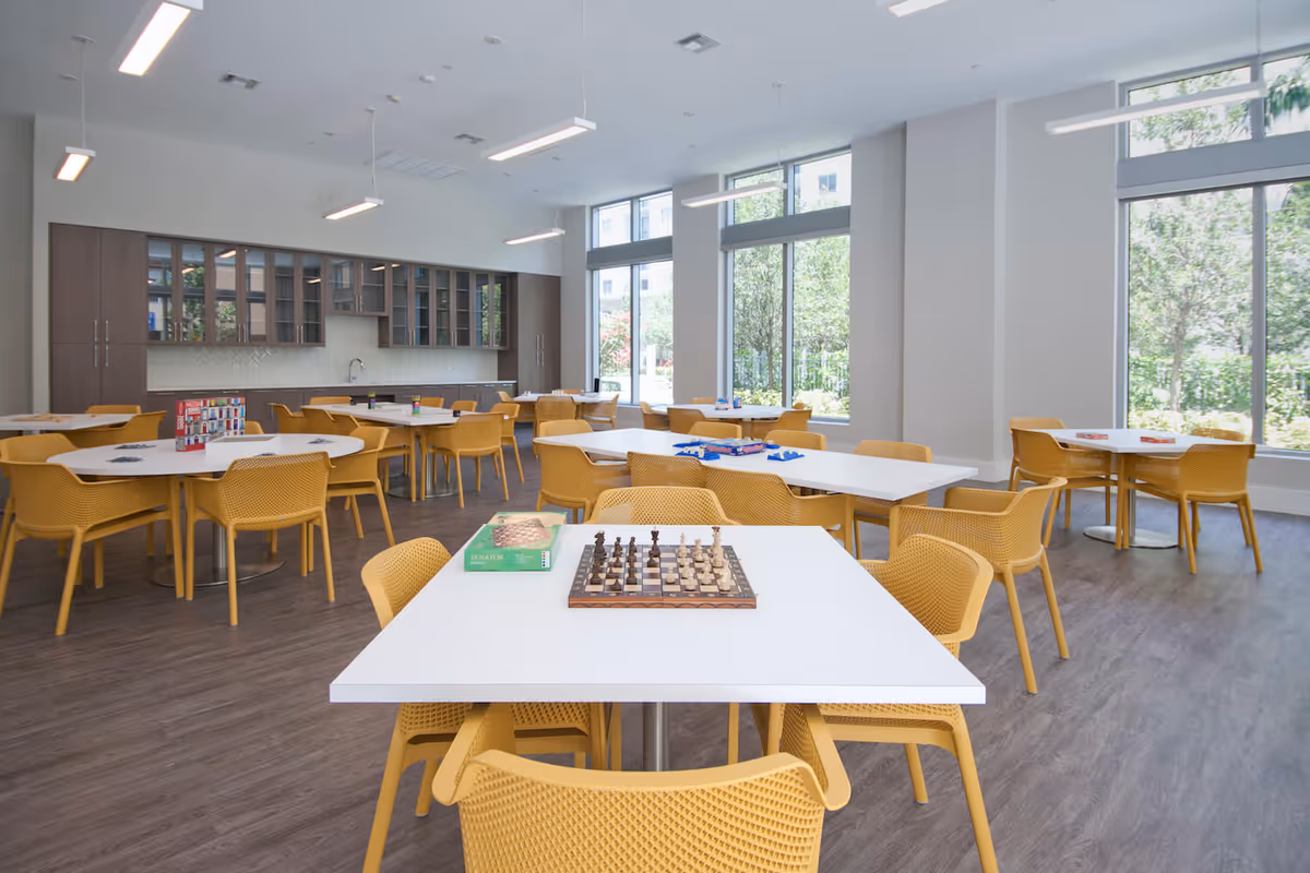 A bright and spacious activity room with large windows letting in natural light. The room has several white tables surrounded by yellow chairs. On the tables are various board games including chess, Connect 4, and other games. The back wall features cabinets with glass doors and a countertop with a sink.