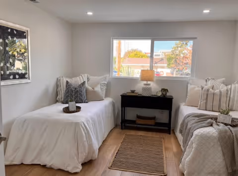 A bright bedroom with two single beds on either side of a black nightstand with a lamp and small decorative items. Each bed is neatly made with white bedding and multiple pillows. A window above the nightstand lets in natural light, and a framed picture hangs on the left wall. The floor is wooden with a small rug between the beds.