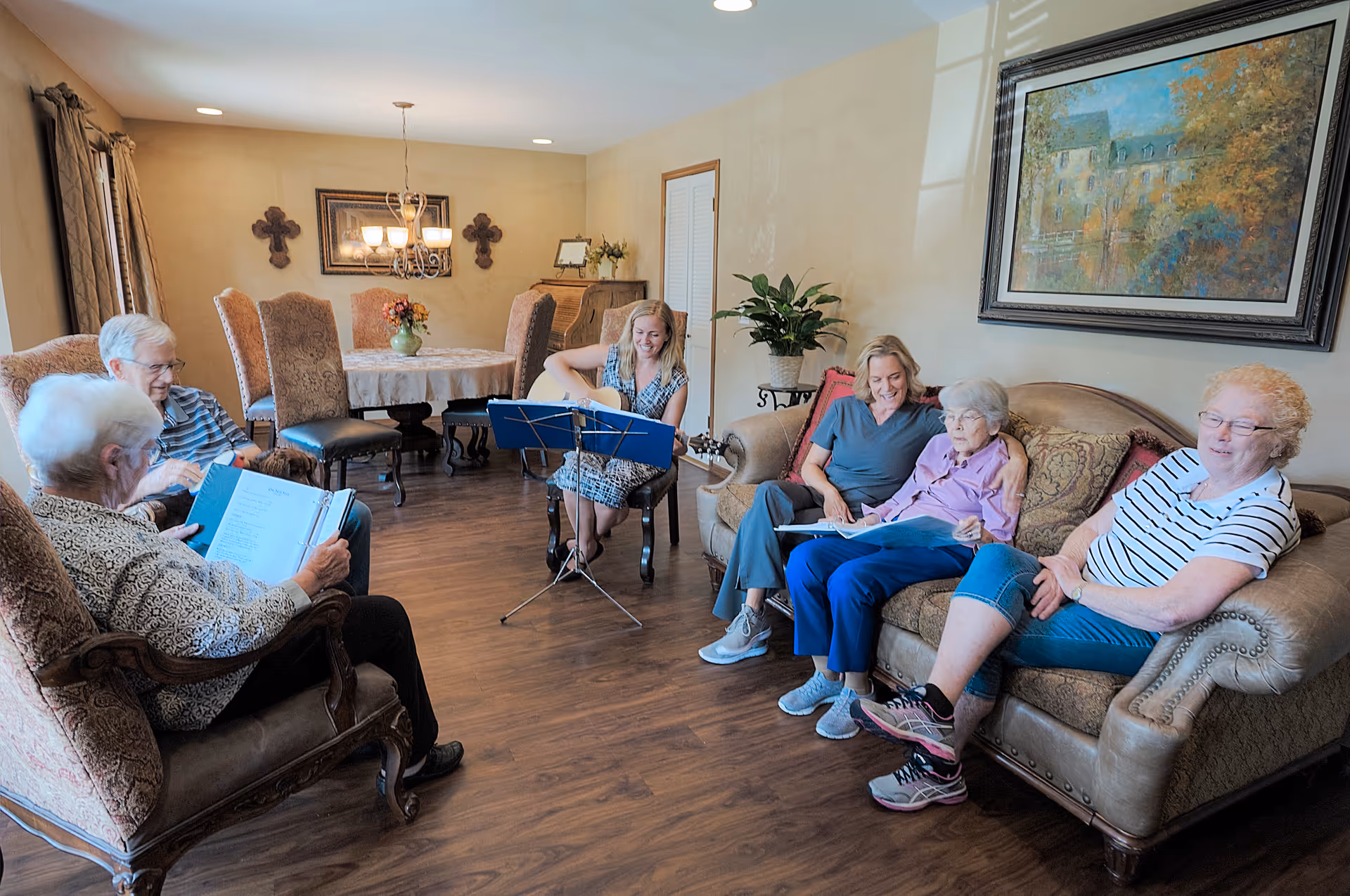 A group of elderly people and a caregiver sitting in a cozy living room. One person is playing a guitar while others are holding songbooks, engaging in a music activity. The room has wooden flooring, a dining table with chairs in the background, wall decorations including crosses and a framed painting, and a potted plant near the sofa.